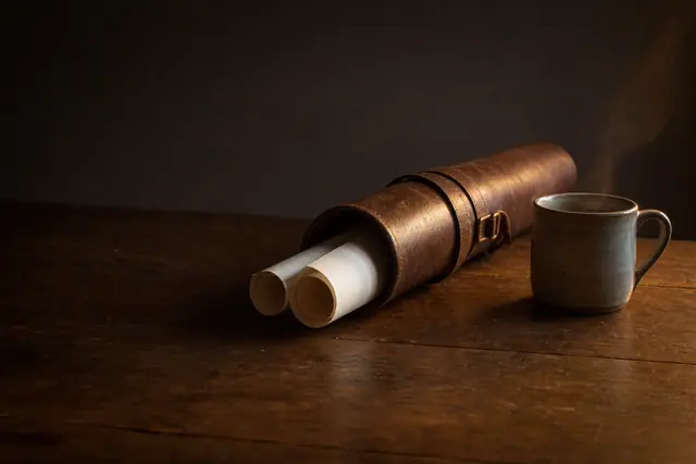 Leather document case with rolled papers beside a steaming ceramic mug on a wooden table.