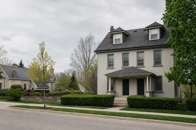 A grey rendered two-and-a-half-storey rental house viewed from across the street on an overcast spring day.