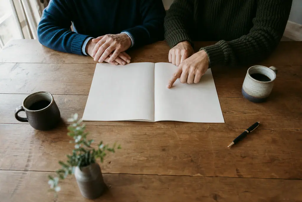 Two people seated side by side at a wooden table, hands resting on an open notebook between two mugs of coffee.