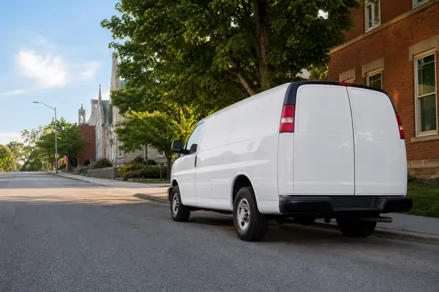 A white cargo van parked on a residential street of brick buildings, with a church spire in the distance.