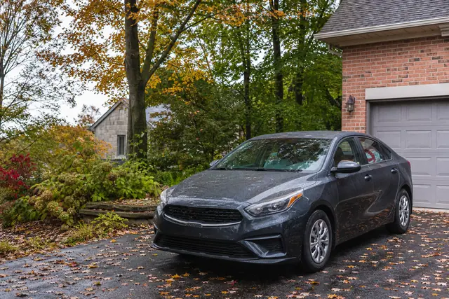 A grey four-door sedan parked on a residential driveway beside a brick house, with autumn leaves across the lawn.