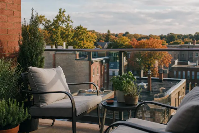 Balcony seating with potted herbs and glass railing overlooking brick buildings and autumn neighbourhood trees at sunset.