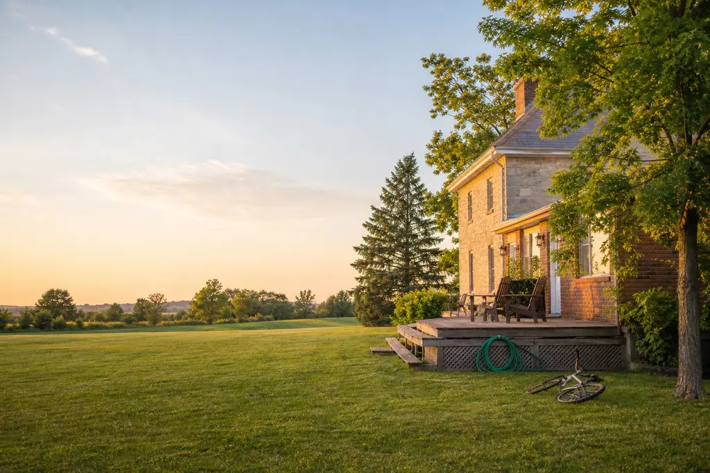 A stone farmhouse at sunset, with a wooden deck, patio chairs and a bicycle lying on the lawn, beside a wide open field.