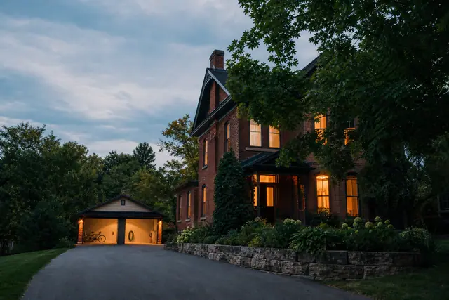 Brick house at dusk with warm lights, detached lit garage, bicycle, driveway, trees, and stone retaining wall.