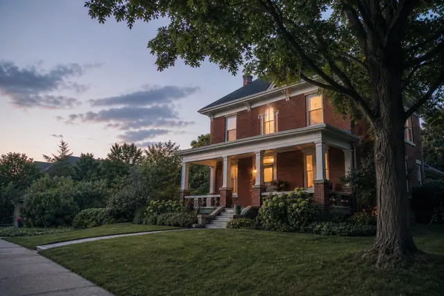A two-storey red-brick house with a lit front porch at dusk, set behind a mown lawn and a mature shade tree.