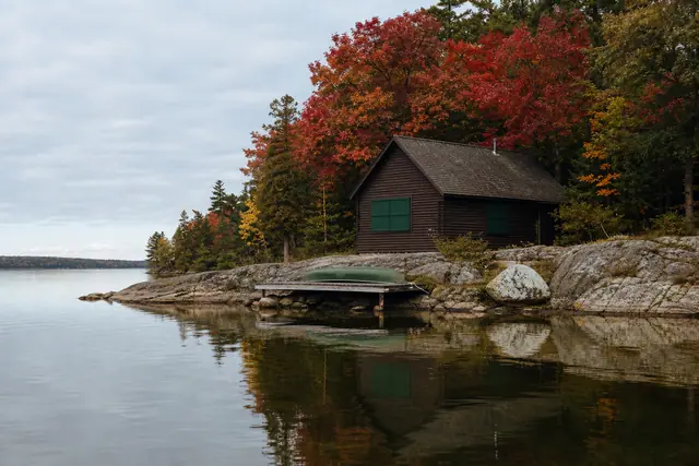 Wooden lakeside cabin with green shutters, upturned green boat on dock, autumn trees reflected in calm water.