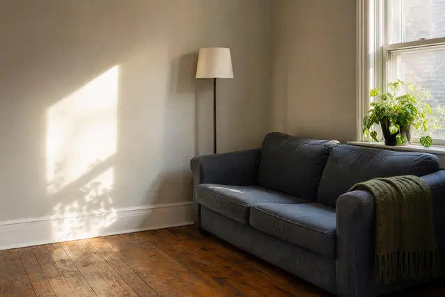 A rented living room corner with a grey sofa, a floor lamp, and afternoon light falling across a bare wall and hardwood floor.
