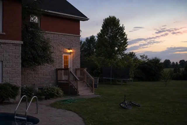 Backyard at dusk with porch light over door, wooden steps, trampoline, bicycle, garden hose, pool ladder.
